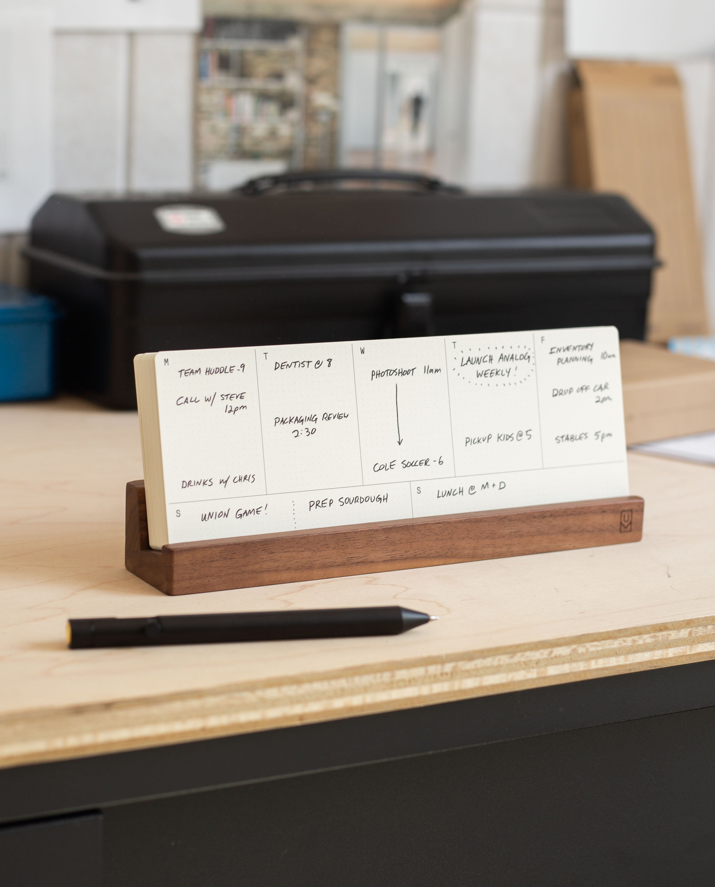 An Analog Annual Kit (Walnut) sits on a desk, displaying handwritten notes and tasks on Daily and Weekly Cards supported by its elegant wooden stand. A black pen lies in front, while a sleek Walnut Card Holder keeps the cards organized. In the background, a black toolbox and other items appear softly blurred.