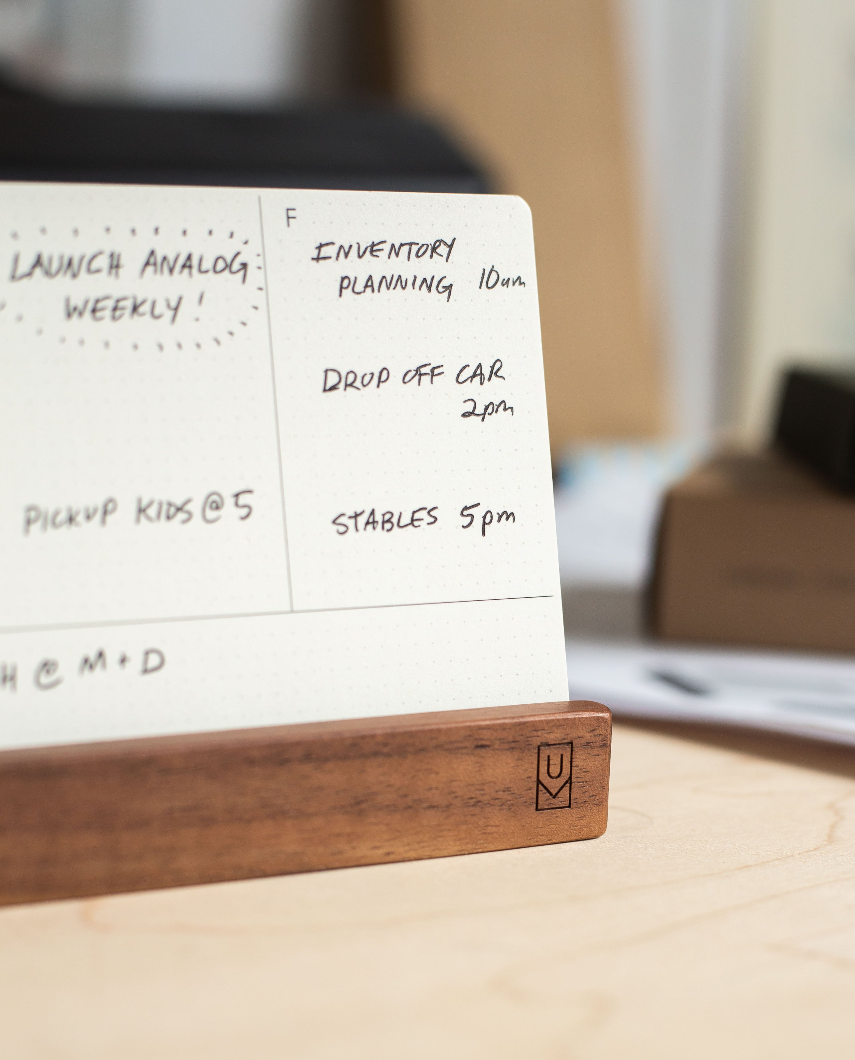 A close-up of a planner on a wooden stand, likely the Analog Weekly Planning Kit (Walnut) card holder, shows handwritten notes for Inventory Planning at 10am, Drop Off Car at 2pm, and Stables at 5pm. Blurred in the background are weekly planner cards from the Analog Weekly Planning Kit (Walnut).