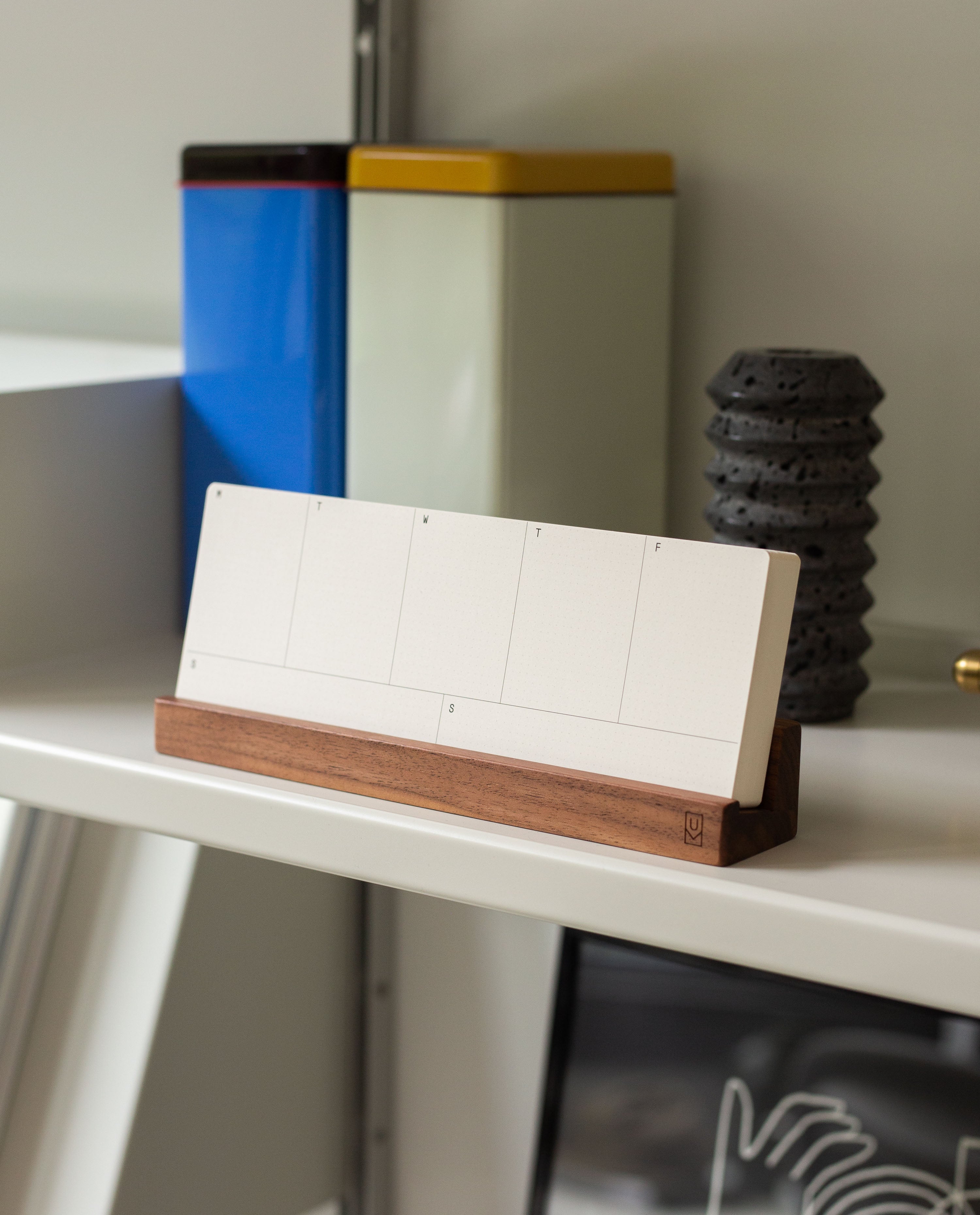 A blank desk calendar from the Analog Weekly Planning Kit (Walnut), featuring a wooden base and walnut card holder, rests on a white shelf alongside colorful tins and a textured black object in the background—an ideal arrangement for organizing your days with its included weekly planner cards.