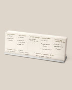 The Analog Weekly Planning Kit (Silver Aluminum) is a white meal planner with handwritten daily notes, featuring dishes like shrimp risotto and tacos, displayed on a silver aluminum stand—ideal for organizing weekly reminders and meals at a glance.
