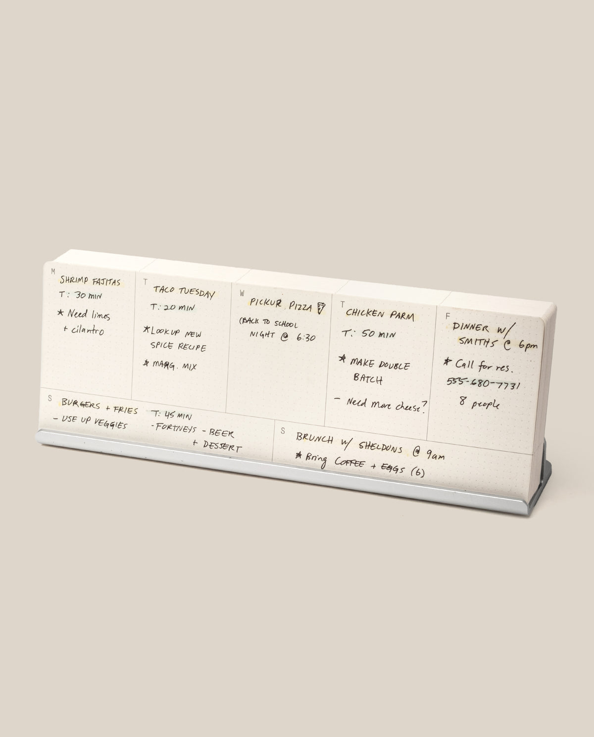 The Analog Weekly Planning Kit (Silver Aluminum) is a white meal planner with handwritten daily notes, featuring dishes like shrimp risotto and tacos, displayed on a silver aluminum stand—ideal for organizing weekly reminders and meals at a glance.