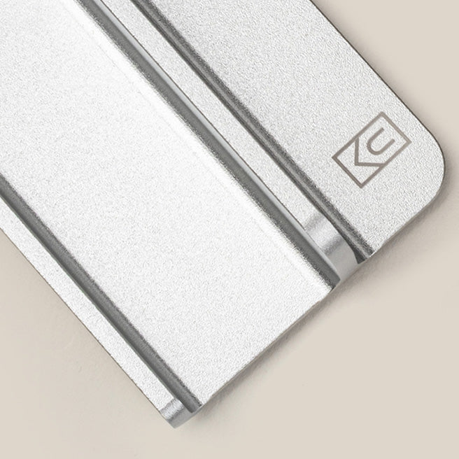 A close-up of a silver metallic object with a smooth, brushed finish and an engraved geometric logo in the top right corner, set against a beige background.