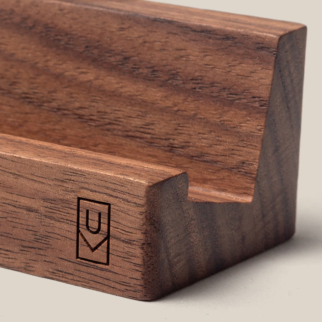 A close-up of a wooden phone or tablet stand with a minimalist design and a geometric logo engraved on the front corner. The wood grain is visible and smooth.