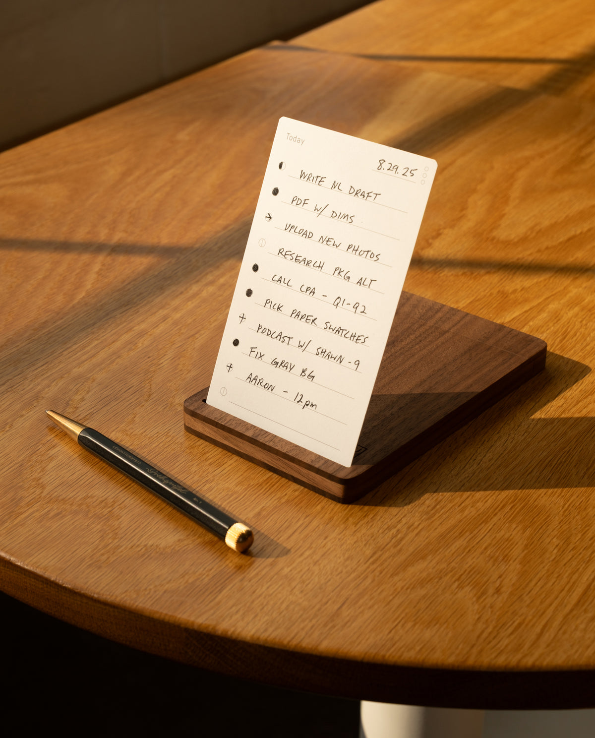 A handwritten to-do list sits on the sunlit wooden table in a walnut card holder beside a refillable pen—tasks like writing a draft and attending a 1 pm meeting are neatly organized in The Analog Bundle (Walnut).