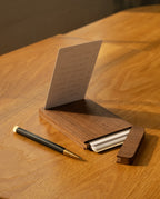 A sleek black and gold pen rests beside the Analog Travel Case + 3 Card Packs (Walnut), which displays a notecard with handwritten notes, all arranged on a light wooden desk bathed in warm sunlight.
