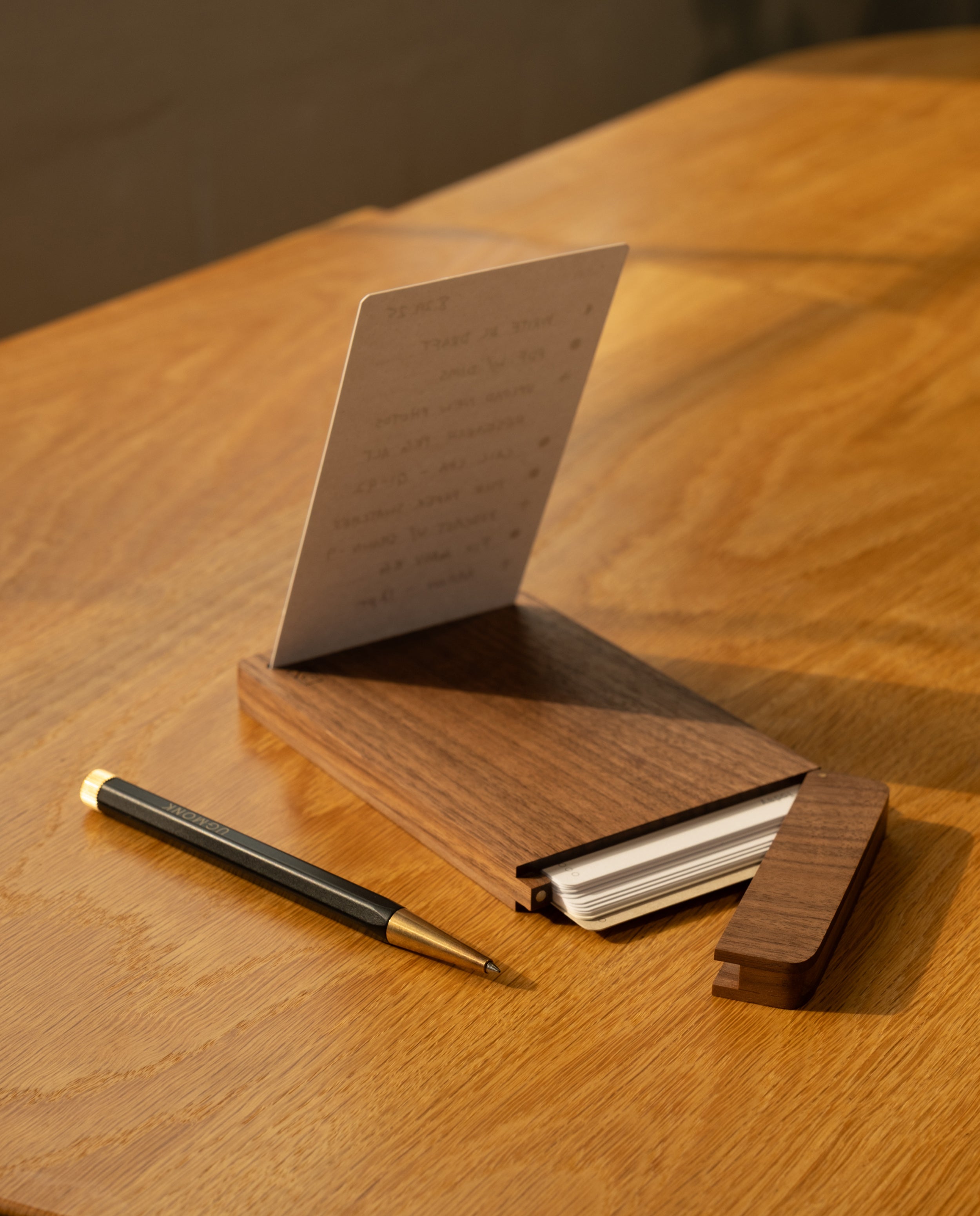 A sleek black and gold pen rests beside the Analog Travel Case + 3 Card Packs (Walnut), which displays a notecard with handwritten notes, all arranged on a light wooden desk bathed in warm sunlight.