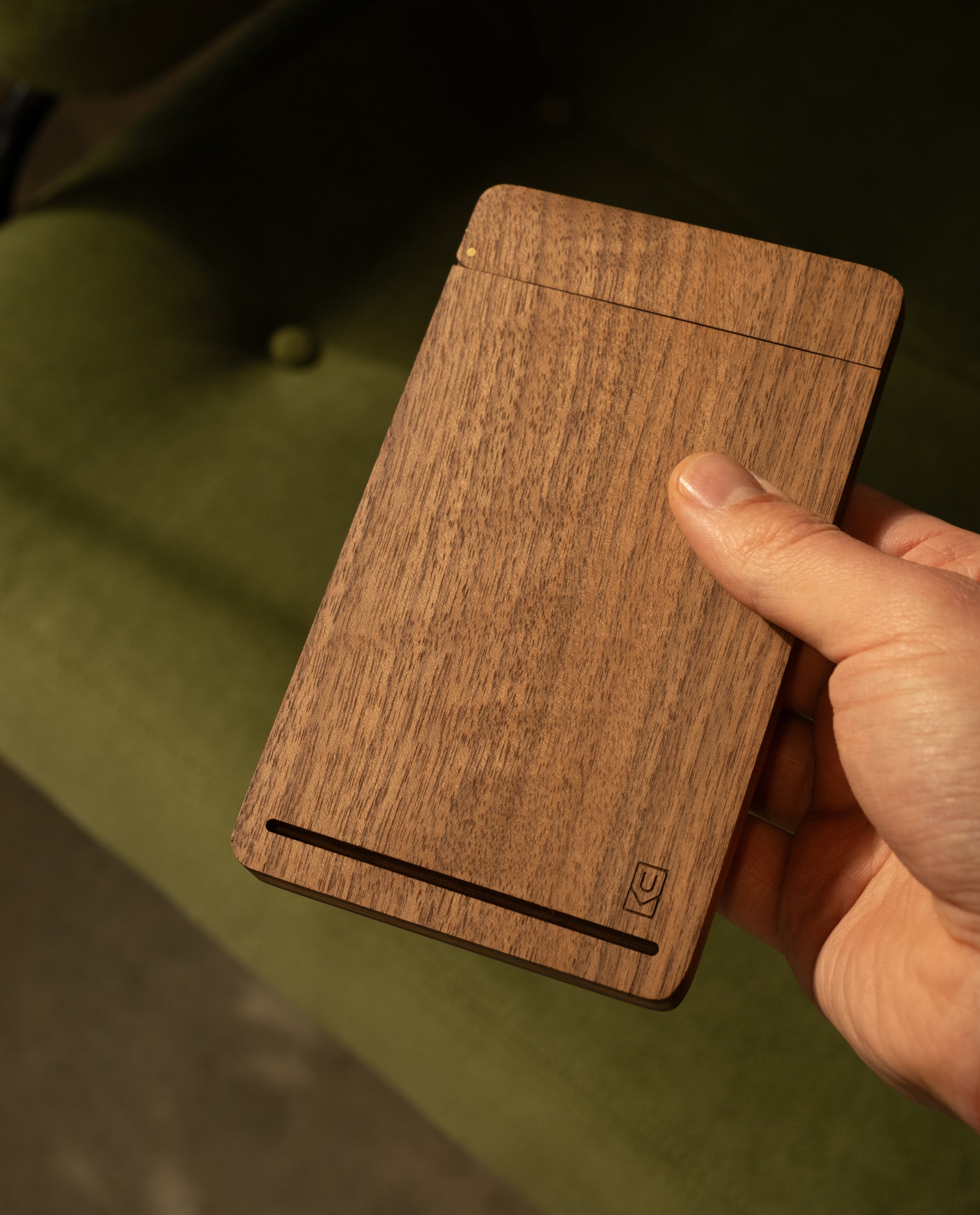 A hand holds a slim, rectangular Analog Travel Case + 3 Card Packs (Walnut) featuring a minimalist design and subtle logo, set against a green fabric background.