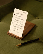 A handwritten to-do list on a small notepad, supported by the premium walnut holder from the Analog Travel Case + 3 Card Packs (Walnut), rests on a green surface. In front of the notepad lies a sleek black and gold pen. The list, featuring tasks like writing, calls, and meetings, evokes the classic task cards included in the Analog Travel Case set.