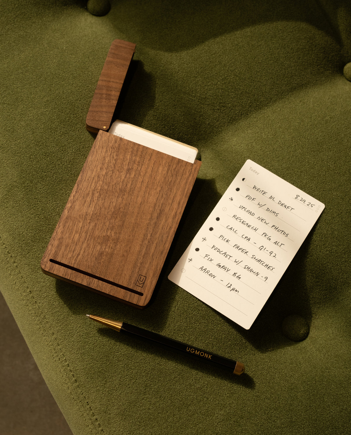 The Analog Bundle (Walnut)—a wooden kit with notecards, an open lid, and a refillable black and gold UGMONK pen—rests on a green cushion. A notecard lists tasks with "$27.25" at the top right corner.