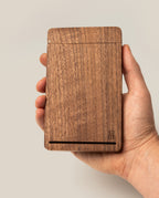 A hand holding the Analog Travel Case + 3 Card Packs (Walnut), a rectangular case crafted from premium walnut with a smooth surface, featuring a thin slot near the bottom and a small logo in the lower right corner—ideal for storing task cards or using as an elegant travel organizer.