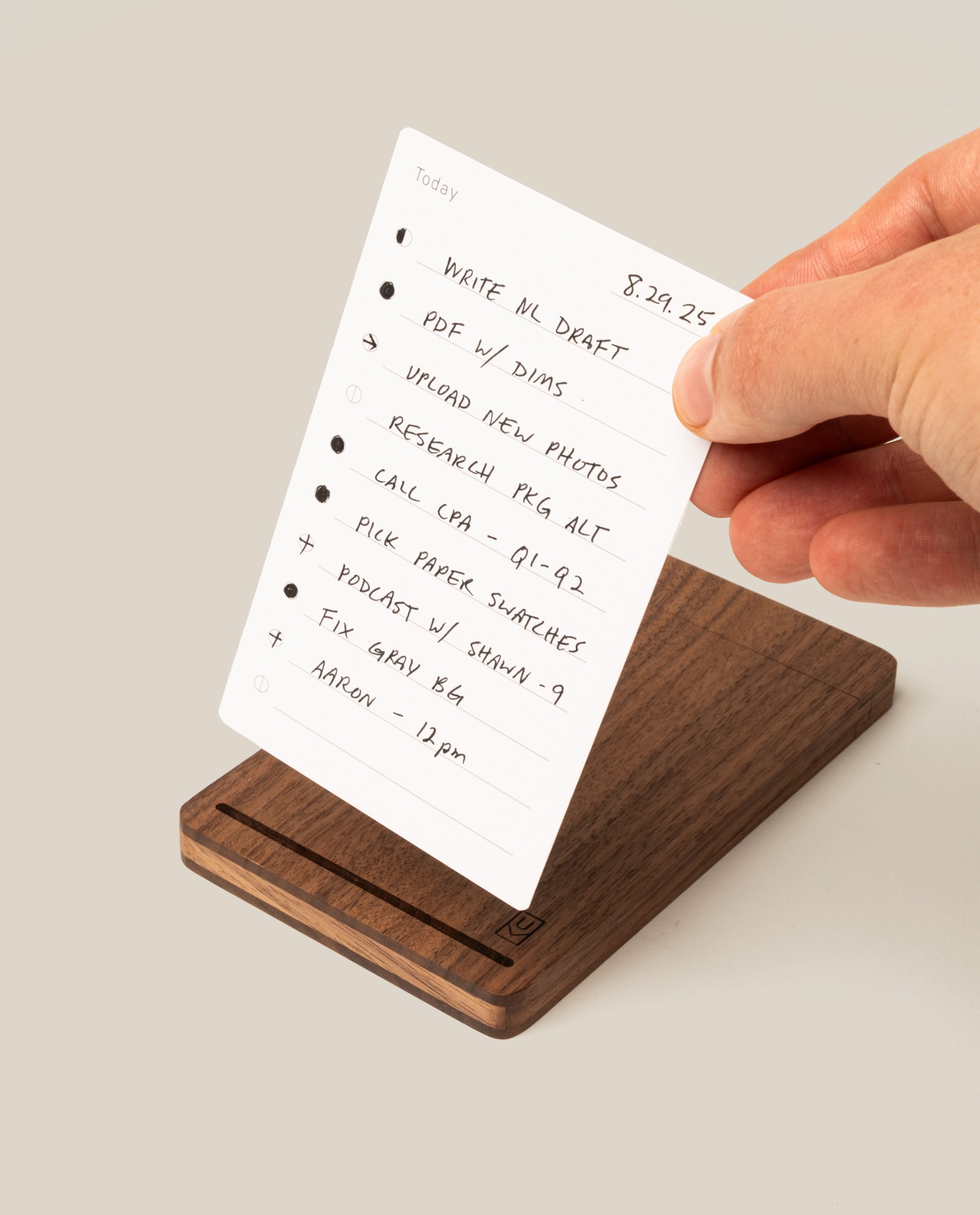 A hand holds a white notecard listing tasks above The Analog Bundle (Walnut), a sleek card holder. The to-do list includes writing, uploading photos, research, calls, and meetings. The scene has a clean, light background.