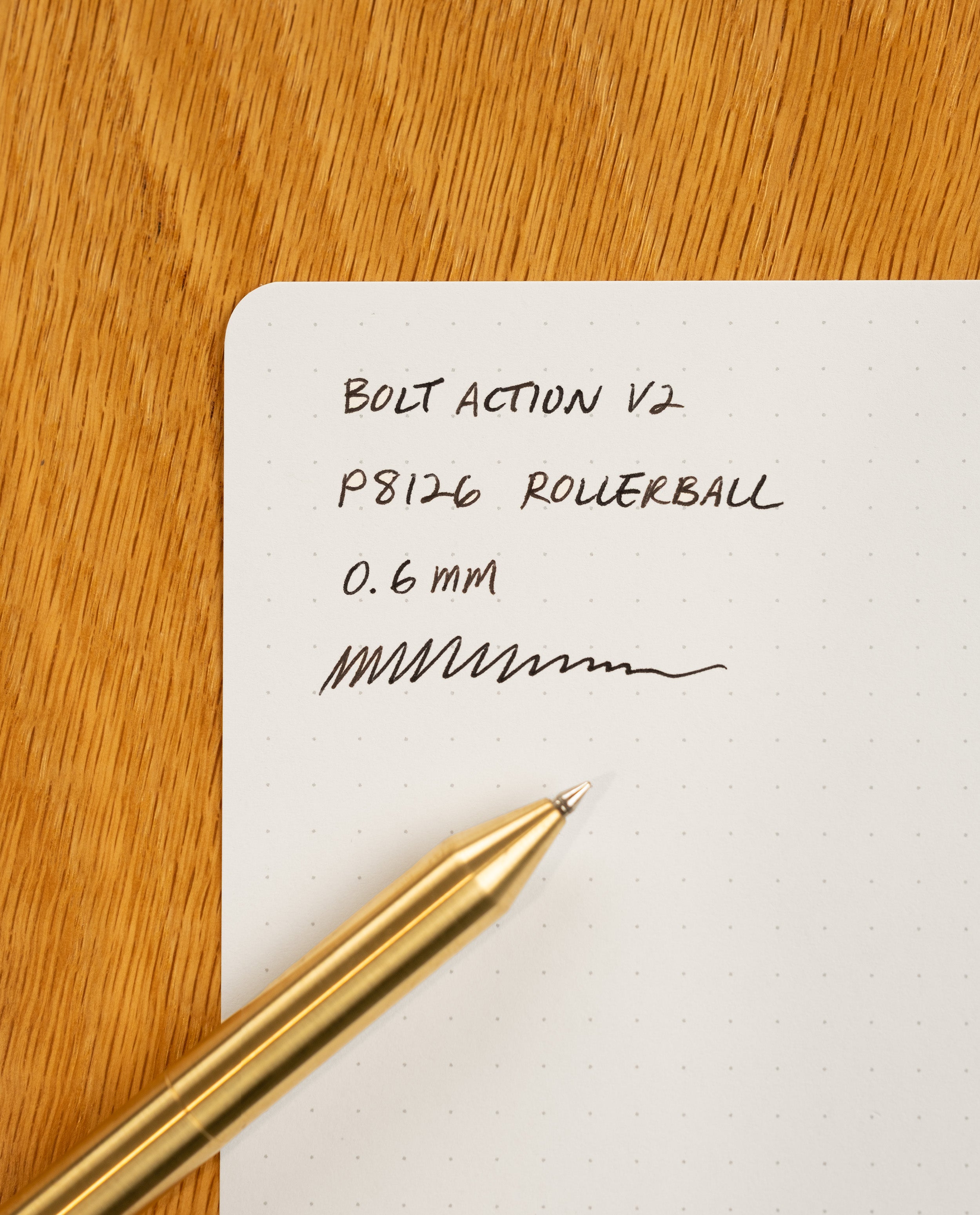 An INVENTERY Bolt Action Pen (Brass) rests on dotted white paper with handwritten notes that read: BOLT ACTION V2 P8126 ROLLERBALL 0.6 mm and some squiggly lines. The paper is placed on a wooden surface.