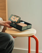 A person holds the Gather 2x2 Base Plate (Black) on a small round wooden table, featuring a phone displaying 4:24, a wooden section, and other compartments seamlessly designed to accommodate magnetic Gather modules on the sturdy powder-coated steel base.