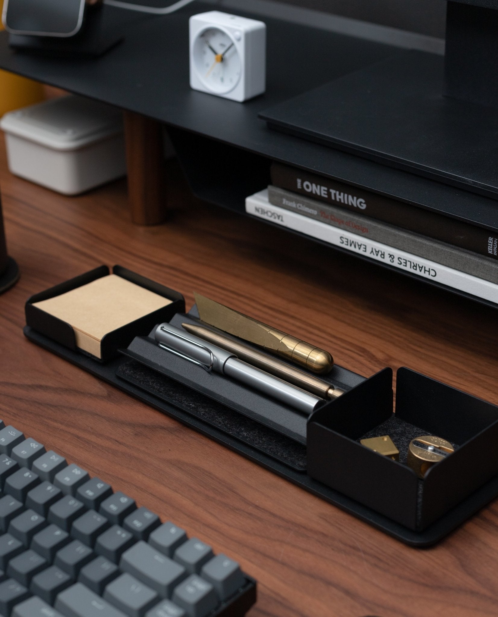 A tidy desk with a keyboard, books, a white clock, and the Gather Note Tray (1x1 - Black) holding pens, a gold utility knife, sticky notes, and a pencil sharpener on a wooden surface.