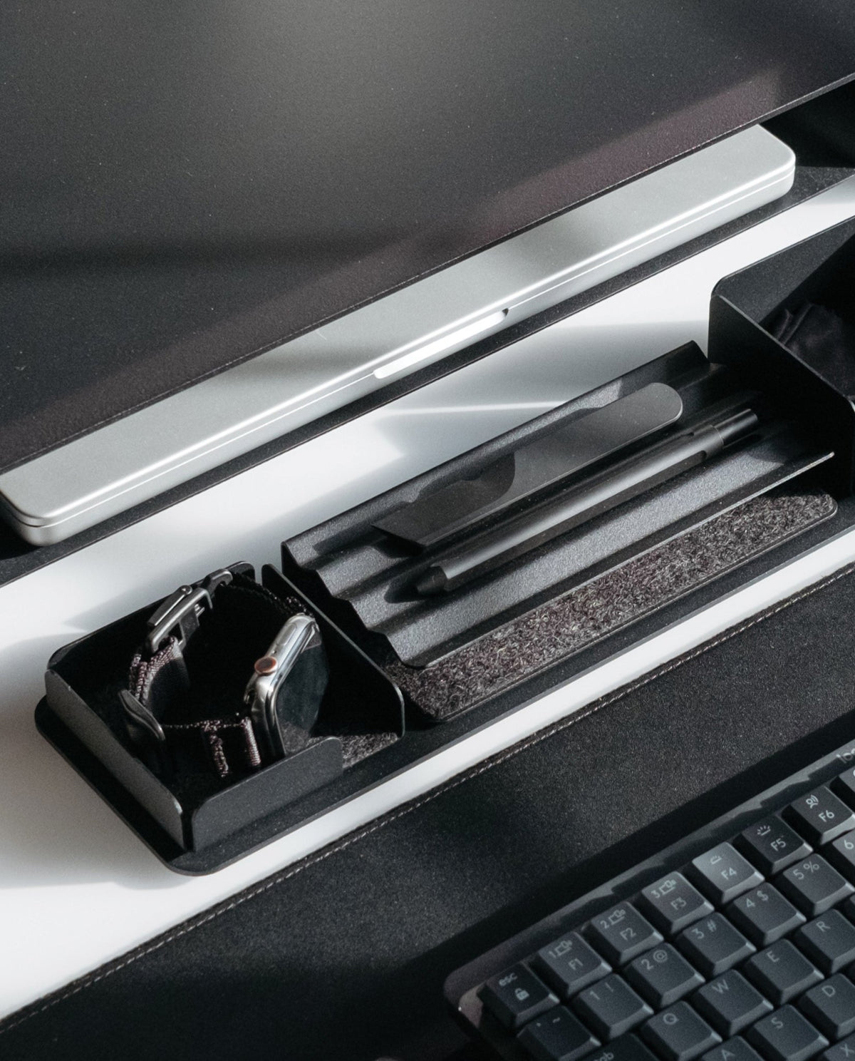 A close-up of a minimalist desktop setup featuring the Gather 1x4 Base Plate (Black) as an organizing tray, paired with a wireless keyboard, monitor, and smartwatch in a tidy, monochrome arrangement.