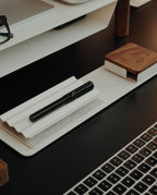 A black pen rests on the Gather 1x4 Base Plate (White) beside a small wooden box and a computer keyboard on a modern, organized desk, complemented by sleek magnetic Gather modules crafted from powder-coated steel.