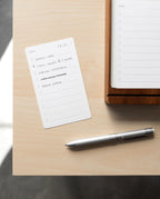 A small notepad with a handwritten to-do list lies on a light wooden desk next to a Craft Design Technology Multifunctional Pen and a larger notepad. The list includes tasks like "Email Josh" and "Call Laura @ 2:30 PM.