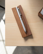 The Craft Design Technology Multifunctional Pen in silver sits on a rectangular wooden holder atop a light wood desk, with soft sunlight casting gentle shadows across the surface.