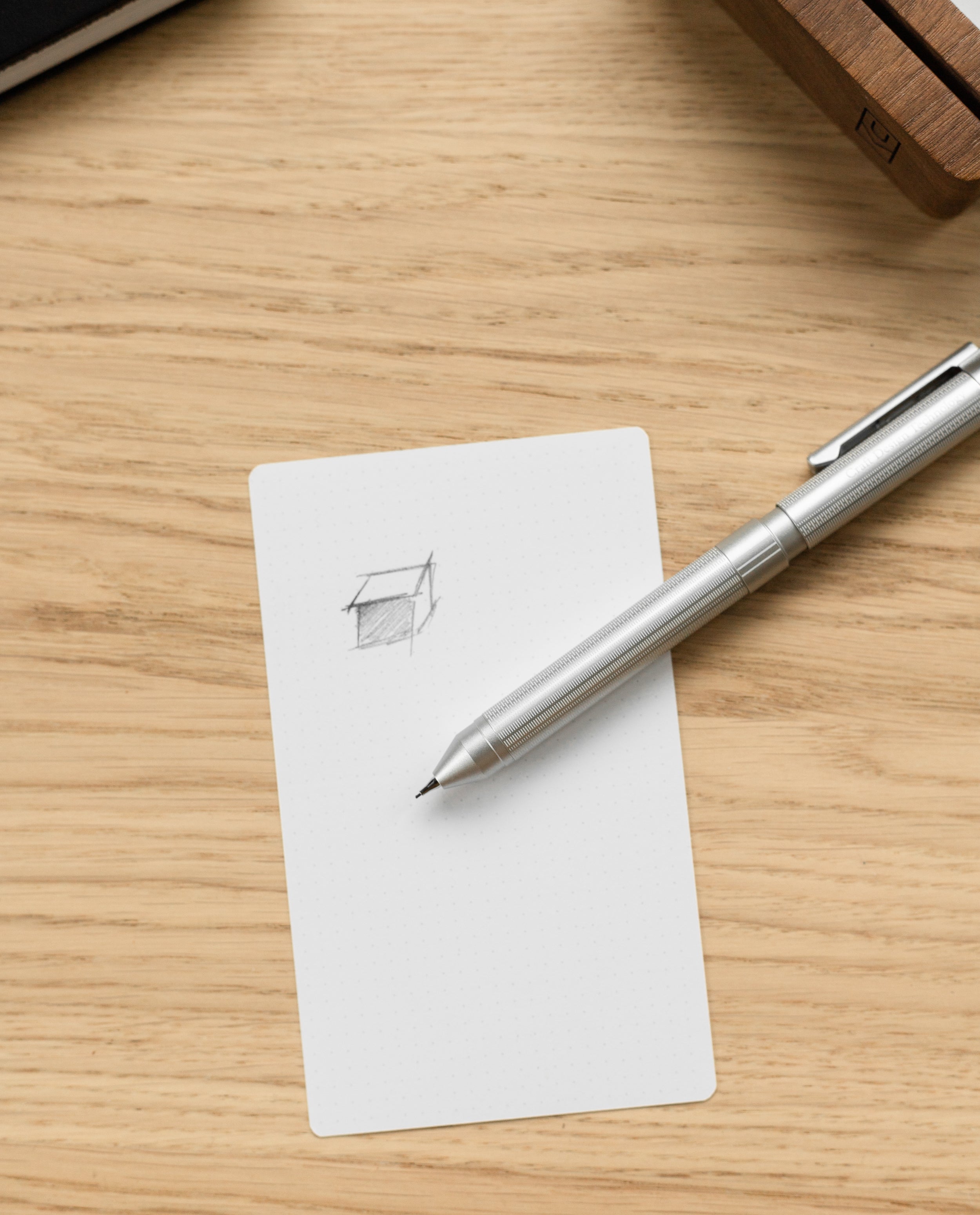 The Craft Design Technology Multifunctional Pen rests on a white notecard featuring a simple pencil sketch of a 3D box, all set atop a light wooden surface.