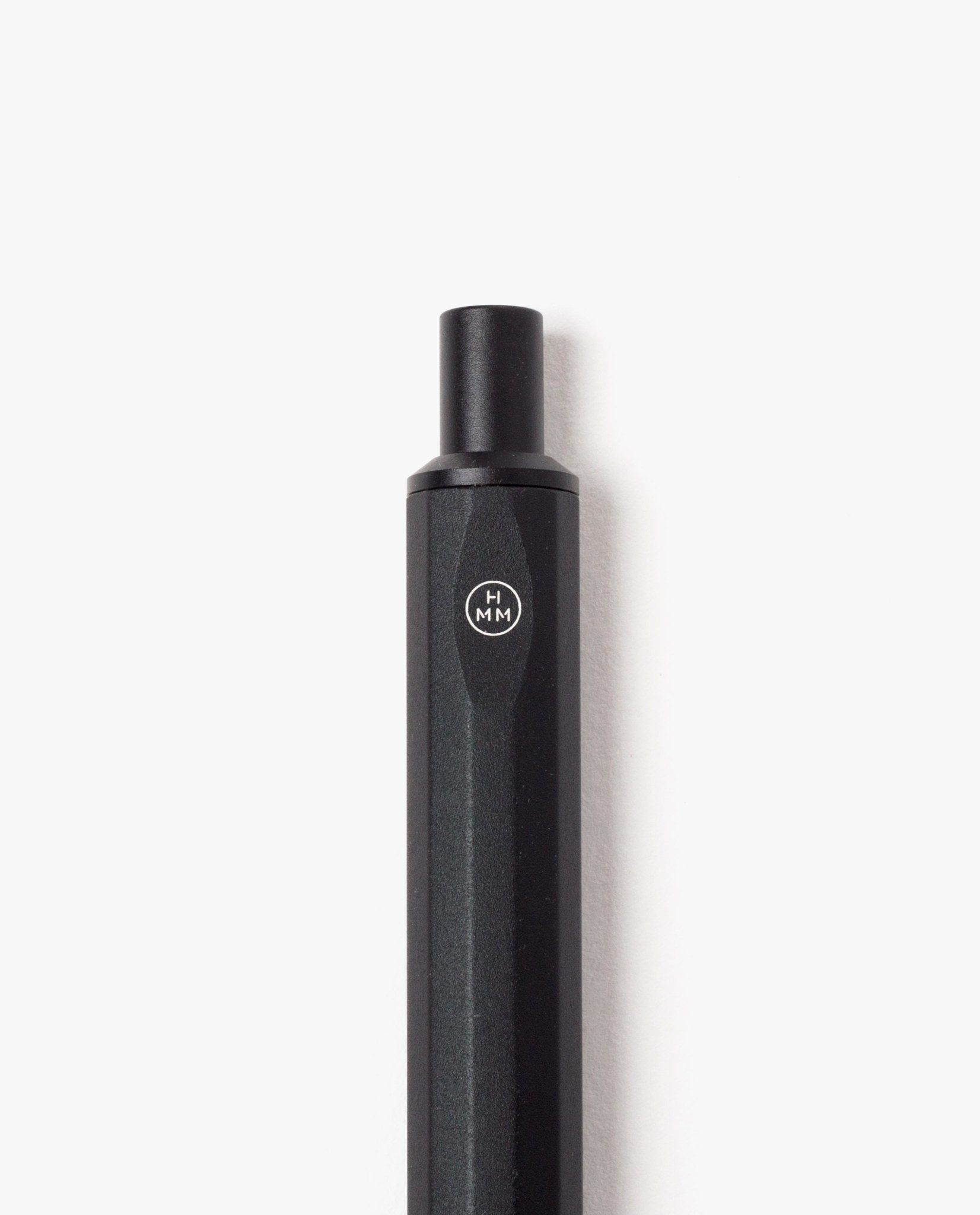A close-up of the HMM Ballpoint Pen (Black)—a hexagonal, black aluminum pen displaying the HMM logo in a circle near the top—photographed against a white background. This elegantly designed pen features a refillable mechanism for long-lasting use.