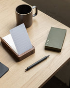 A HMM Ballpoint Pen (Black), a notepad in a wooden holder, a green box, and a ceramic mug sit on a light wooden desk, creating a minimal, organized workspace.