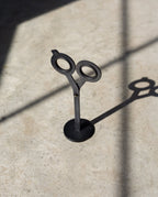 A pair of HMM Scissors (Black) stands upright on a magnetic metal base, casting a distinct shadow on a sunlit surface with intersecting lines of light and shadow.