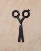 HMM Scissors (Black) with round handles and a minimalist design rest closed on a light wood surface, handles up. A magnetic metal base complements their sleek look.