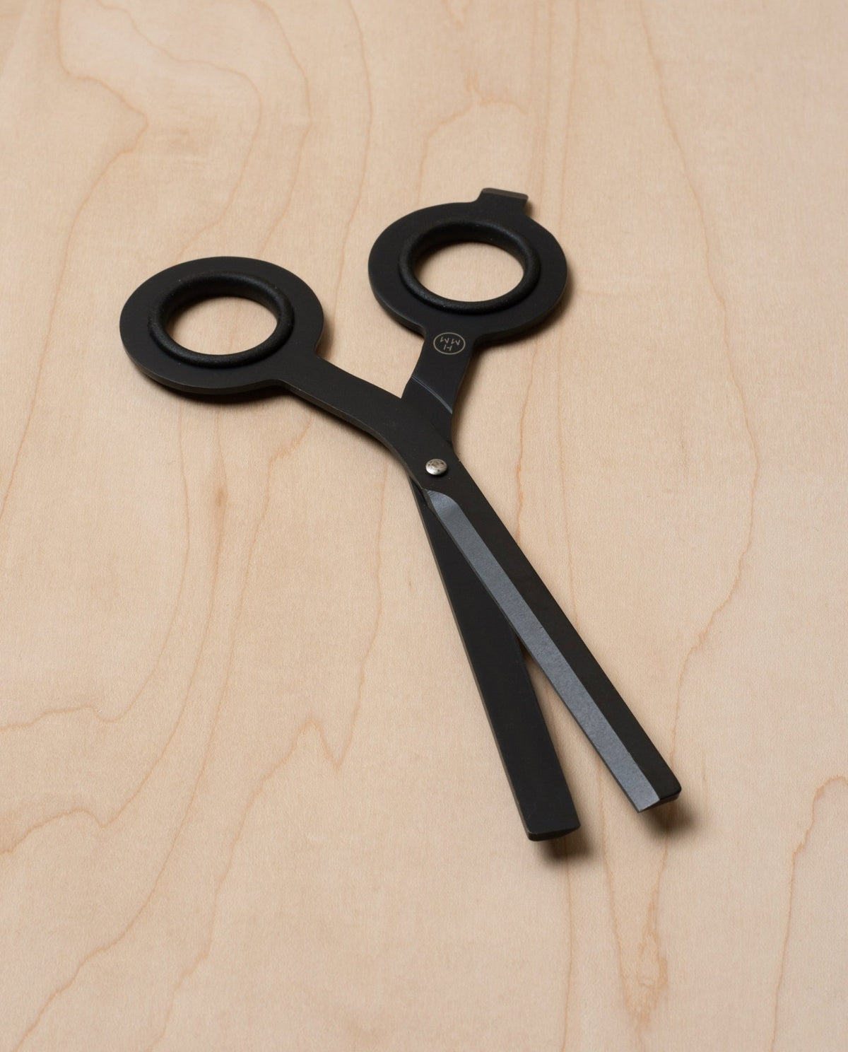 The HMM Scissors (Black) feature round handles and two blunt, flat blades. Decorative and non-functional, they rest on a light wood surface and can be displayed on a magnetic metal base.