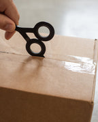 A hand uses HMM Scissors (Black), a small circular cutting tool with a built-in box cutter, to slice through clear tape sealing a brown cardboard box.