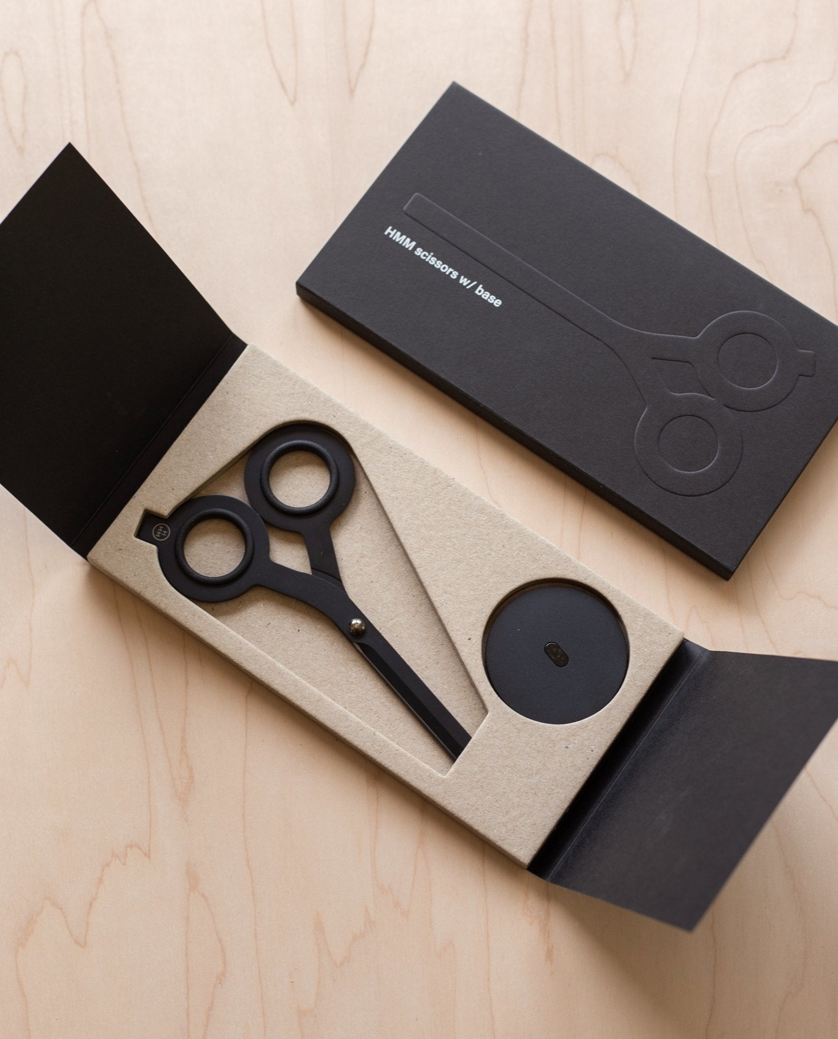 A pair of HMM Scissors (Black) with round handles is shown in an open black box featuring minimalist packaging and a magnetic base, while a similar closed box rests nearby on a light wood surface.