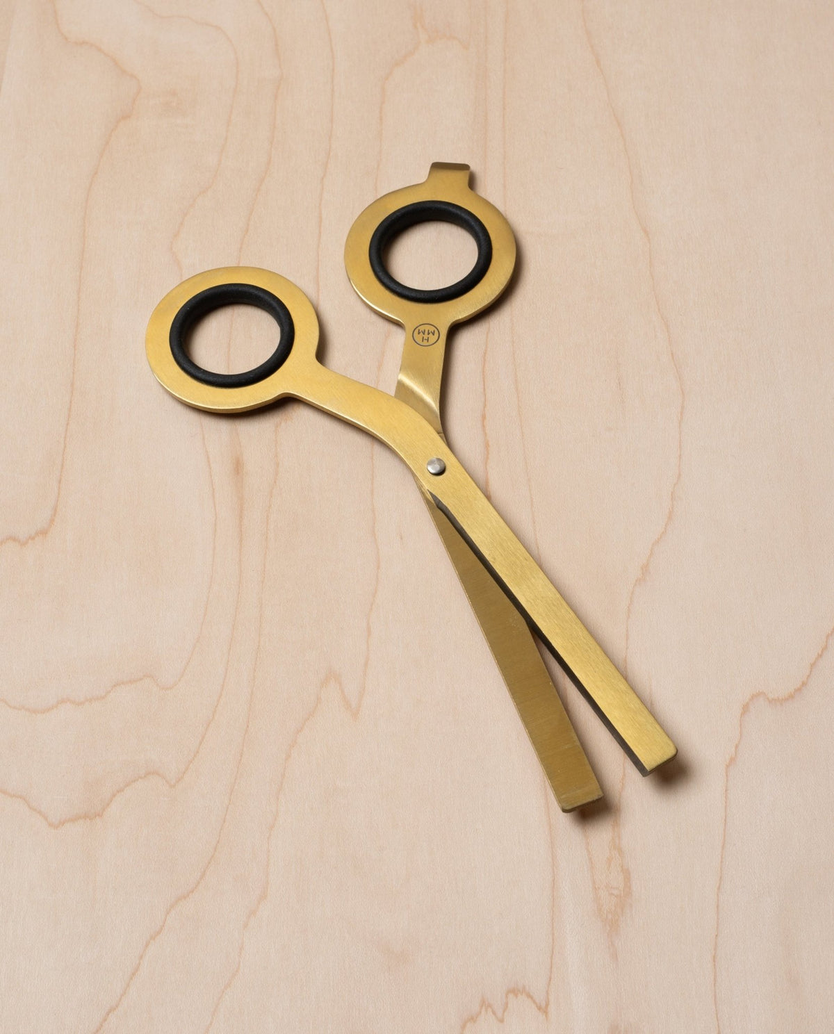 The HMM Scissors (Gold) with round black finger holes rest closed on a light wooden surface, positioned diagonally across the image.