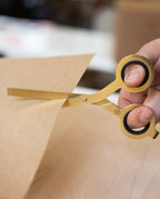 A hand holding a pair of sleek HMM Scissors (Gold) is cutting a piece of brown paper on a wooden surface.