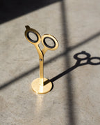 A pair of HMM Scissors (Gold) stands upright on a metal base, casting a distinct shadow on a sunlit concrete floor.