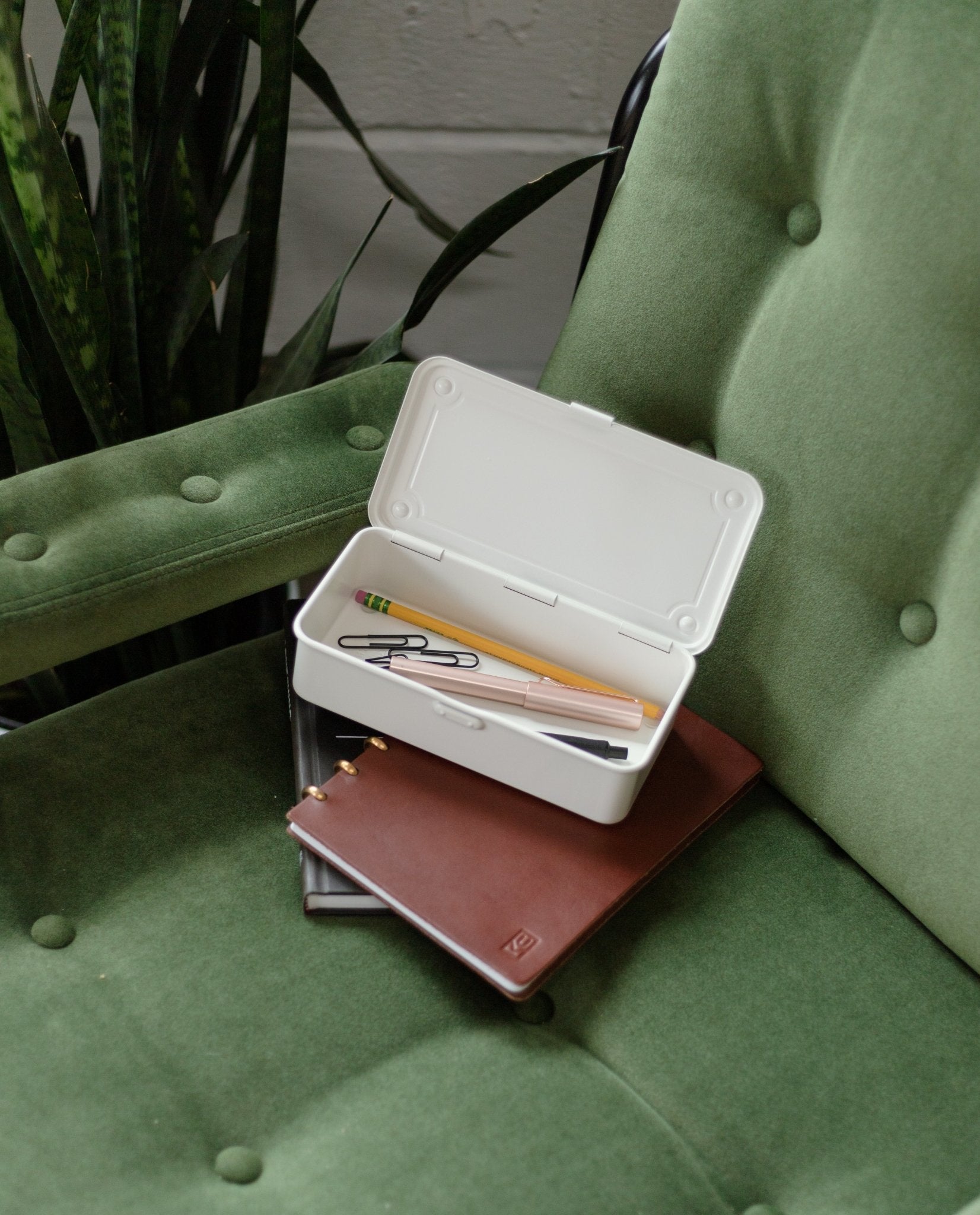 A Toyo Steel Stackable Storage Box T-190 (White), designed in Japan, sits open on a green cushioned chair holding pens, pencils, a paperclip, and erasers inside. Nearby are two notebooks, and a leafy plant can be seen beside the chair.
