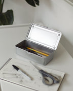 A Toyo Steel Stackable Storage Box T-190 (Silver) with its lid open sits on a white desk, holding a pencil and pen. In front are an open sketchbook, black marker, and scissors. A plant is in the background. Made in Japan.