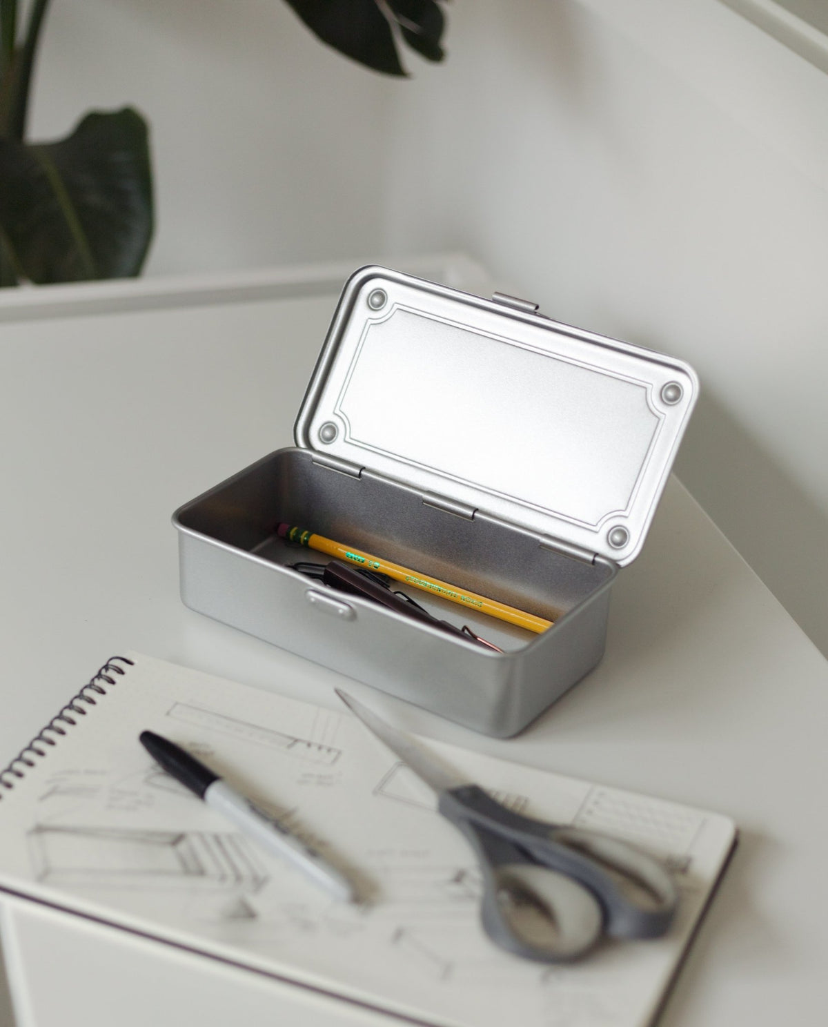 A Toyo Steel Stackable Storage Box T-190 (Silver) with its lid open sits on a white desk, holding a pencil and pen. In front are an open sketchbook, black marker, and scissors. A plant is in the background. Made in Japan.