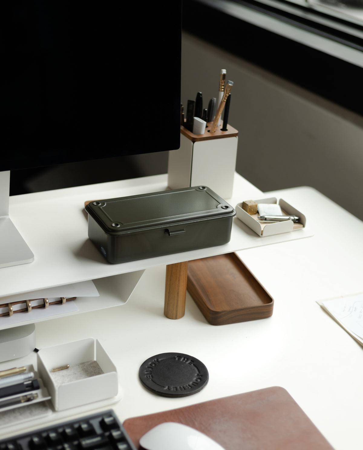 A modern desk features a computer, pen holder, trays with office supplies, and a Toyo Steel Stackable Storage Box T-190 (Military Green) made in Japan, all arranged near a large window that provides natural light.