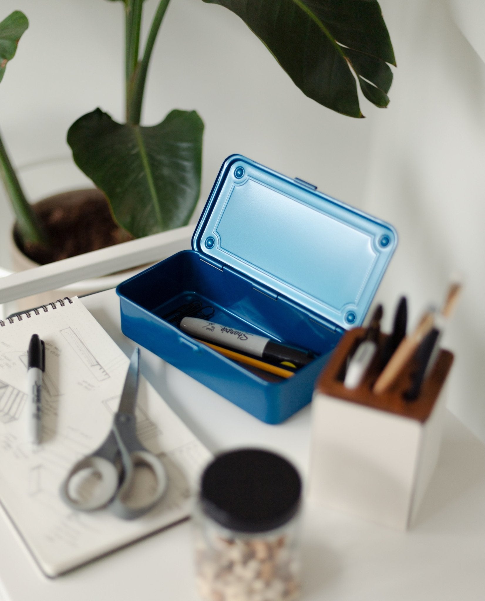 A blue Toyo Steel Stackable Storage Box T-190 sits open, holding markers inside and evoking the iconic style of classic Japanese toolboxes. Next to it on the white desk are a sketchbook, scissors, pens organized in a holder, a small jar, and a green plant—all thoughtfully arranged to inspire creativity.