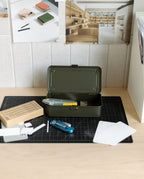 A Toyo Steel Stackable Storage Box T-190 (Military Green), made in Japan, sits open on a desk with a utility knife, Swiss army knife, cardboard box, and notecards. Above the workspace, mood board photos and materials are pinned to the wall.