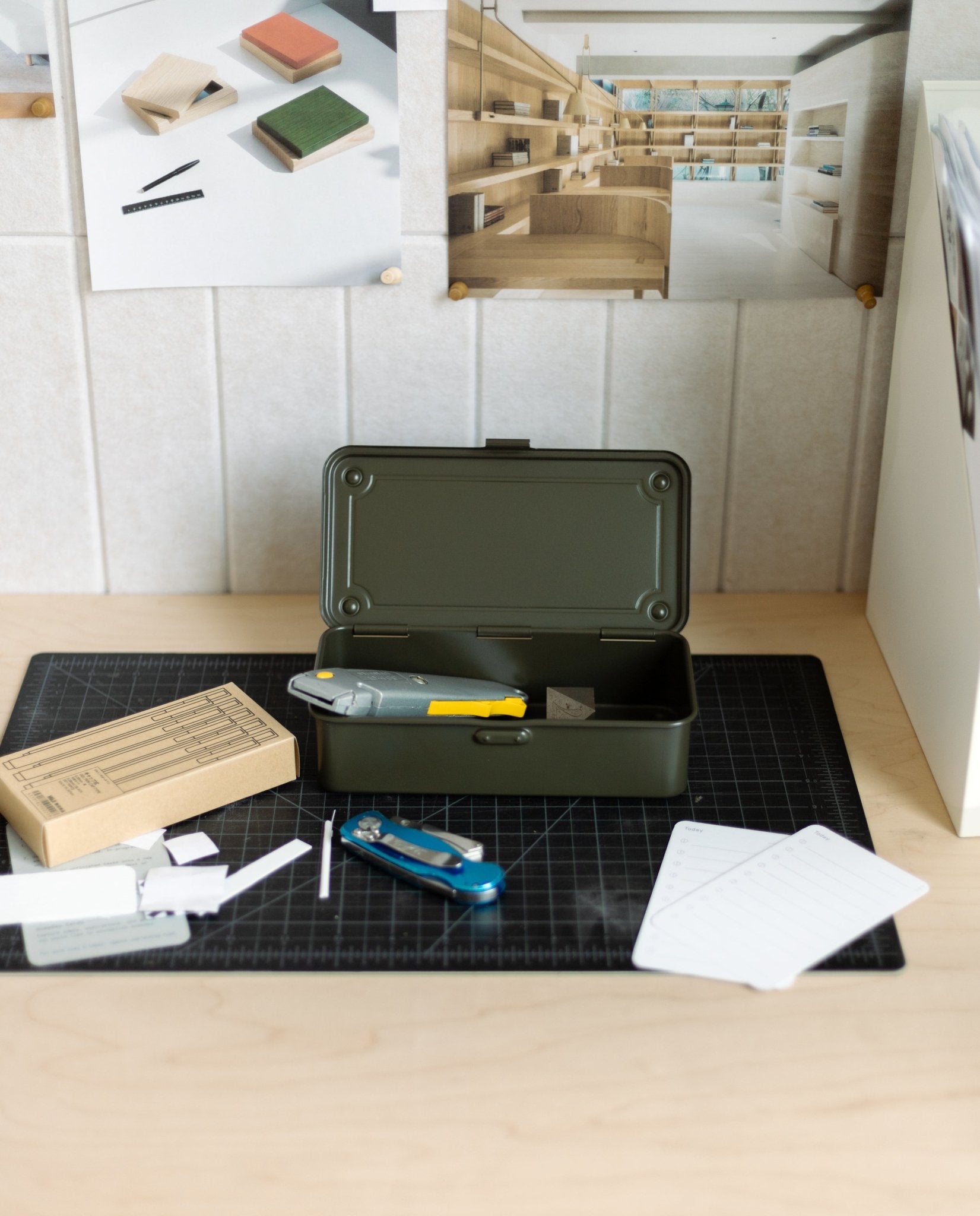 A Toyo Steel Stackable Storage Box T-190 (Military Green), made in Japan, sits open on a desk with a utility knife, Swiss army knife, cardboard box, and notecards. Above the workspace, mood board photos and materials are pinned to the wall.