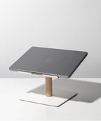A closed gray laptop sits on the Gather Laptop Stand (White/Maple), featuring a minimalist white and maple wood design, placed on a white surface against a light gray background.