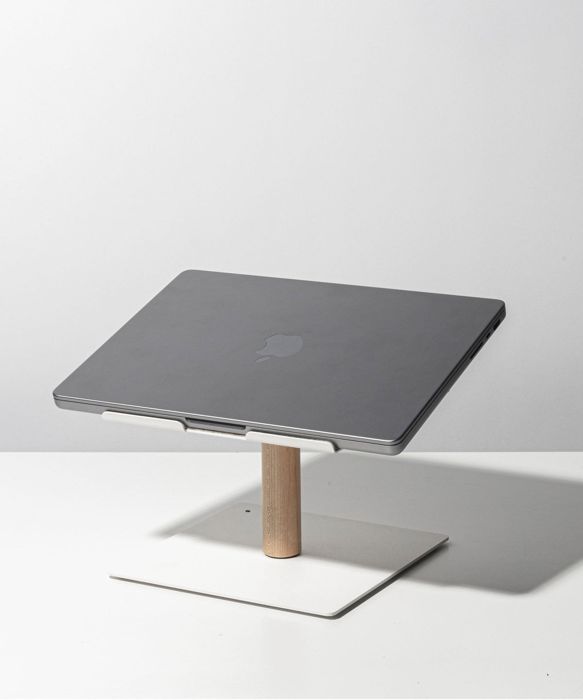 A closed gray laptop sits on the Gather Laptop Stand (White/Maple), featuring a minimalist white and maple wood design, placed on a white surface against a light gray background.