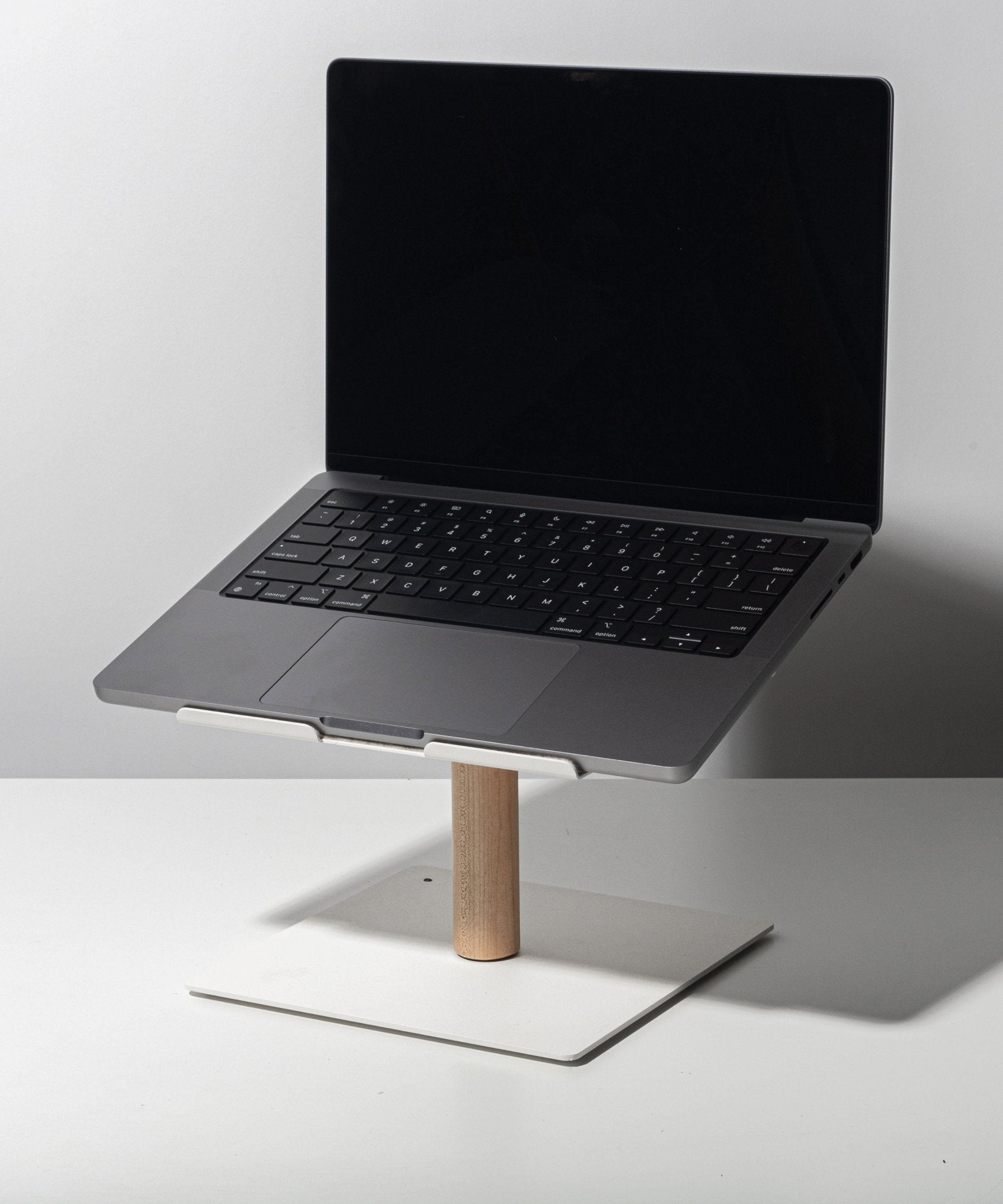 A black-screen laptop rests on the Gather Laptop Stand (White/Maple), featuring a maple rod and white base, placed on a white table against a plain light background.
