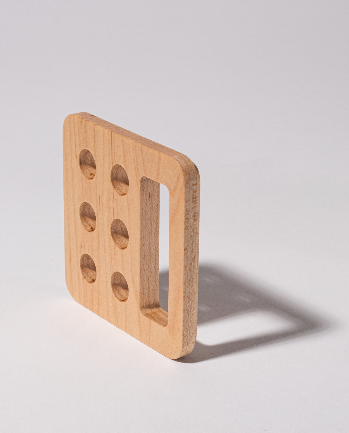 A square maple Gather Pencil Cup Lid, handcrafted in Pennsylvania from solid wood, features five round holes on one side and a large rectangular cutout, casting a shadow on a light surface.