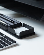 A Gather Note Tray (1x1 - Black) with a pen and a black note holder containing white paper sits on a white desk next to part of a black and white computer keyboard. This sleek organizer is proudly made in the USA, combining functionality with modern style.