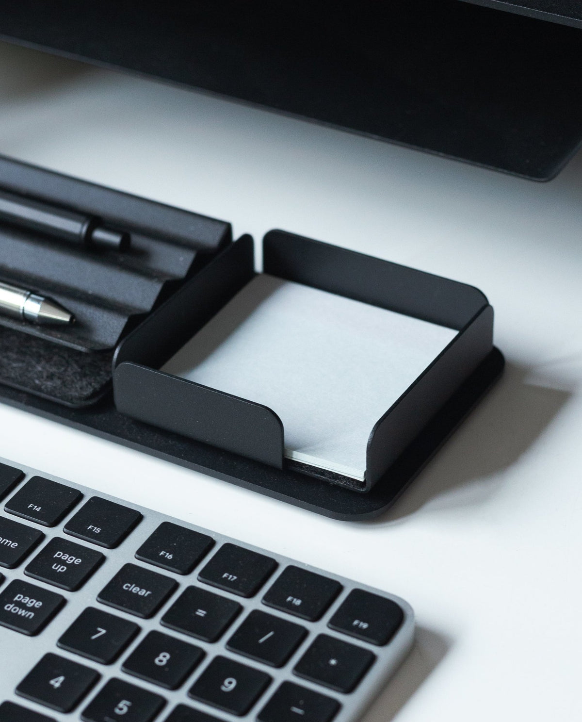 A black keyboard sits next to a sleek black Gather Note Tray (1x1 - Black) containing blank white notepaper, with pens in a tray above it, all on a white desk.