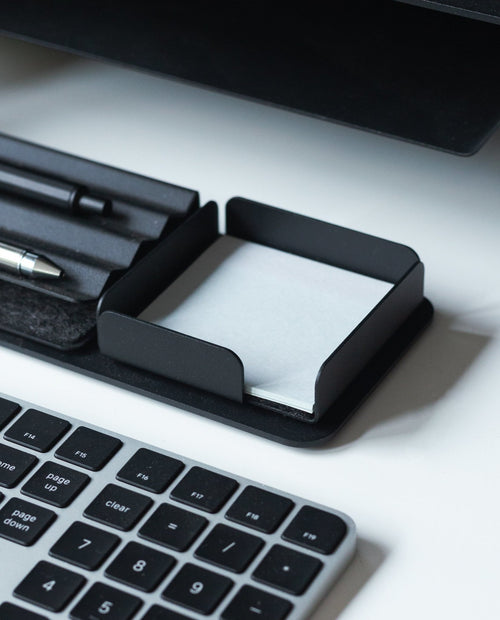 A black keyboard sits next to a sleek black Gather Note Tray (1x1 - Black) containing blank white notepaper, with pens in a tray above it, all on a white desk.