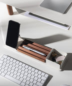 A minimalist white desk setup includes a wireless keyboard, silver pen on a walnut pen holder, smartphone, and the Gather Wood Zigzag Tray (2x1 - Walnut) holding keys and USB drives—all neatly arranged under bright lighting.