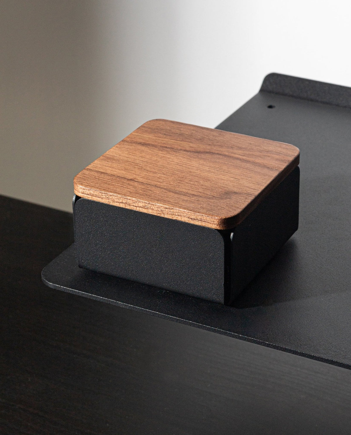 A Gather Solid Wood Lid (Walnut) rests atop a small, square device with a black base, positioned on the edge of a black desk or shelf against a plain background.