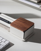 A Gather Solid Wood Lid (Walnut) organizer, featuring a black pen inside and a white base, sits on a white desk near a computer keyboard and stacked books.